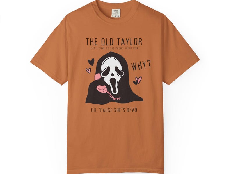 May include: A rust-colored t-shirt featuring a graphic of a ghost-faced figure holding a pink telephone. Text includes "THE OLD TAYLOR" and "WHY?" with hearts. The shirt's design is a playful take on a horror theme.