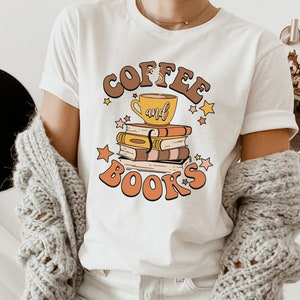 May include: White t-shirt with a retro-style graphic design featuring a coffee cup and books stacked on top of each other. The text reads "Coffee and Books" in a stylized font.