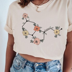 May include: A white t-shirt with a floral design that represents the chemical structure of serotonin. The text "Serotonin" is written in black above the design.
