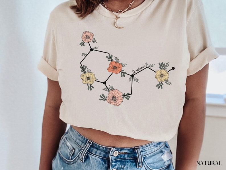 May include: A white t-shirt with a floral design that represents the chemical structure of serotonin. The text "Serotonin" is written in black above the design.