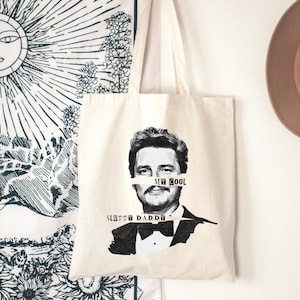 May include: A white canvas tote bag with a black and white image of a man wearing a tuxedo. The text "MR. COOL" is partially obscured by a white tape across the man's face. The text "SLOTH DADDY" is printed below the image.