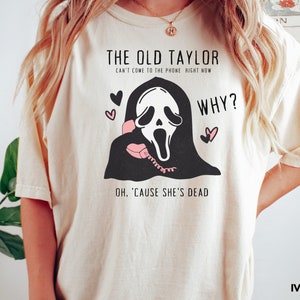 May include: Ivory-colored t-shirt featuring a black and white graphic of a ghost-faced figure holding a pink telephone. Text includes "THE OLD TAYLOR" and "WHY?" with the phrase "OH, 'CAUSE SHE'S DEAD". Two pink hearts are also present.