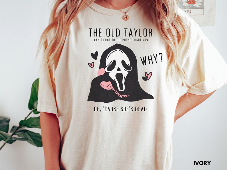 May include: Ivory-colored t-shirt featuring a black and white graphic of a ghost-faced figure holding a pink telephone. Text includes "THE OLD TAYLOR" and "WHY?" with the phrase "OH, 'CAUSE SHE'S DEAD". Two pink hearts are also present.