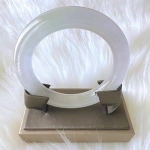 May include: A smooth, translucent, pale lavender and white circular bangle bracelet resting on a taupe-colored rectangular display stand with two curved supports that hold the bracelet upright in a vertical position.