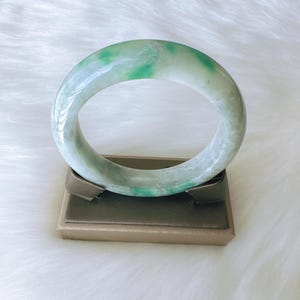 May include: A smooth, polished jade bangle bracelet featuring a translucent white base with vibrant green mottled patterns throughout the circular band. The bangle has a rounded profile and a glossy finish, showcasing natural mineral variations.