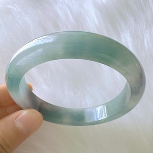 May include: A translucent jade bangle bracelet in shades of green and gray. The smooth, polished surface reflects light, showcasing the natural variations in color. The bracelet is a circular shape, designed to be worn on the wrist.