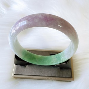 May include: A smooth, polished bangle bracelet featuring a gradient of colors, transitioning from a soft lavender and white tone at the top to a vibrant, mottled green hue at the bottom, showcasing a natural, translucent stone texture.