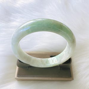 May include: A smooth, polished jade bangle bracelet featuring a translucent appearance with soft gradients of pale green and milky white. The circular stone band has a rounded exterior profile and a polished finish that reflects light across its surface.
