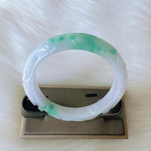 May include: A circular bangle bracelet crafted from translucent white stone with vibrant green inclusions. The surface of the bangle features intricate carved floral patterns that wrap around the entire circumference of the smooth, polished band.