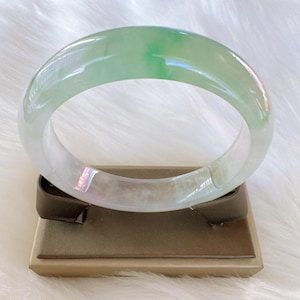 May include: A smooth, polished jade bangle bracelet featuring a translucent appearance with variegated light green and white color patterns. The circular jewelry piece has a rounded profile and a glossy finish, showcasing natural mineral variations.