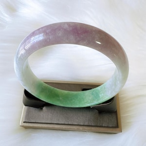May include: A smooth, polished bangle bracelet featuring a gradient of colors, transitioning from a soft lavender and light purple at the top to a vibrant, translucent green at the bottom, with a rounded, circular shape and a glossy, reflective surface.