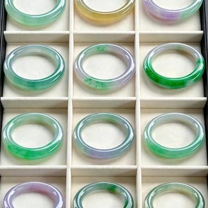 May include: A collection of twelve smooth, circular jade bangles displayed in a black partitioned tray. The bangles feature various translucent shades of green, lavender, and yellow, each with a polished finish and a rounded, continuous band design.