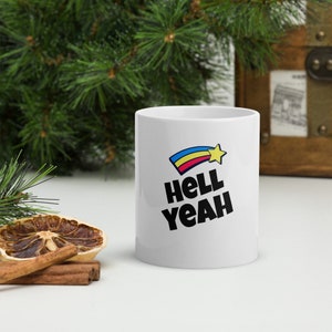 May include: White ceramic mug with a black and white graphic of a rainbow shooting star with a yellow star and the text "Hell Yeah".