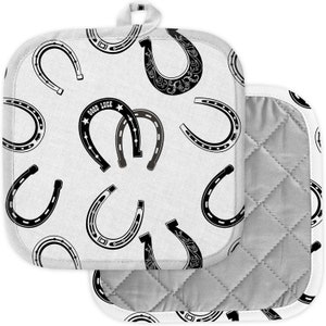 Puede incluir: Dos agarraderas cuadradas blancas con un patrón de herraduras. Las herraduras son negras y tienen varios diseños, incluyendo las palabras "GOOD LUCK" en una. Las agarraderas tienen un interior acolchado gris y un ribete blanco.