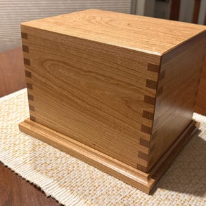 May include: A rectangular wooden box with a light brown finish. The box features dovetail joints at the corners and sits on a slightly raised base. The wood grain is visible on the surface.