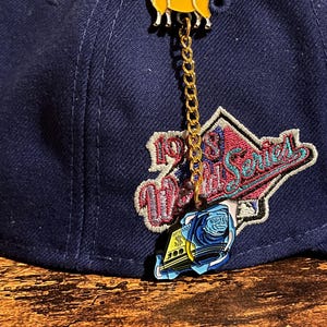 May include: Navy blue baseball cap with a gold chain and two enamel pins. The top pin is a yellow goat, and the bottom pin is a blue and yellow design. The cap has a patch that says "1988 World Series".