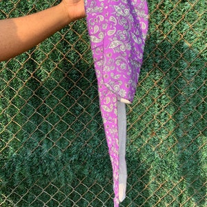 May include: A purple paisley patterned head covering. The fabric features a repeating design of white and light green paisley shapes. The head covering is held up against a chain link fence and green foliage.