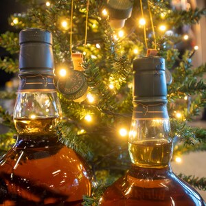 May include: Two brown glass bottles filled with amber liquid, likely liquor, with dark green caps. The bottles are in front of a Christmas tree decorated with lights and bottle cap ornaments.