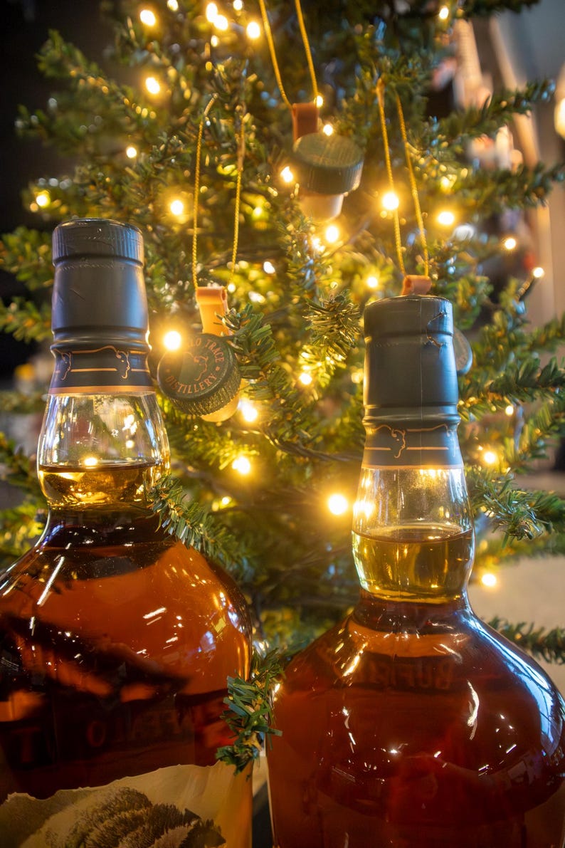 May include: Two brown glass bottles filled with amber liquid, likely liquor, with dark green caps. The bottles are in front of a Christmas tree decorated with lights and bottle cap ornaments.