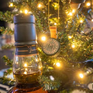 May include: A brown glass bottle of whiskey with a black cap and gold accents, partially filled with amber liquid. Bottle cap ornaments with the text "Buffalo Trace Distillery" hang on a Christmas tree with warm white lights.