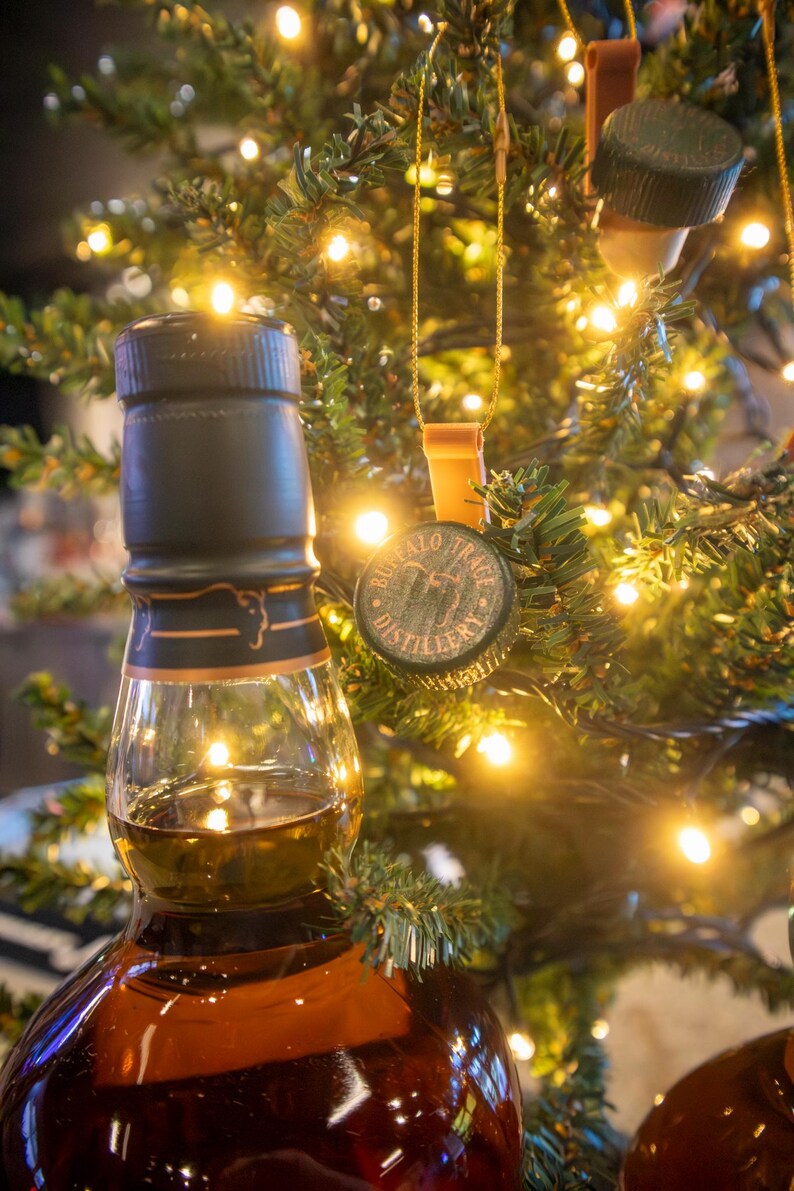 May include: A brown glass bottle of whiskey with a black cap and gold accents, partially filled with amber liquid. Bottle cap ornaments with the text "Buffalo Trace Distillery" hang on a Christmas tree with warm white lights.
