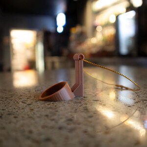 May include: A light brown, 3D-printed wine bottle holder with a ring and a vertical support. A thin, gold-colored wire is attached to the holder. The holder is resting on a speckled, light brown surface.
