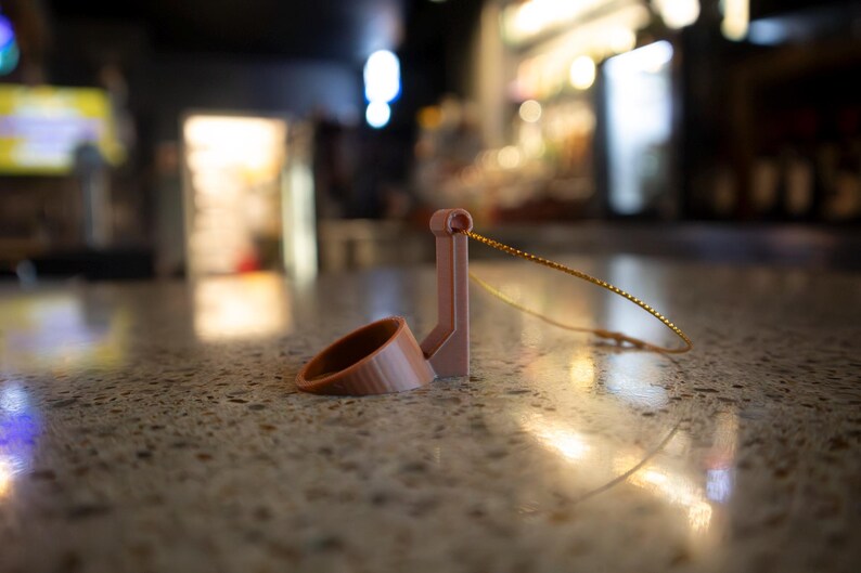 May include: A light brown, 3D-printed wine bottle holder with a ring and a vertical support. A thin, gold-colored wire is attached to the holder. The holder is resting on a speckled, light brown surface.