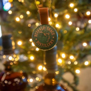 May include: A Buffalo Trace Distillery bottle cap ornament with a brown leather strap and gold-colored string. The green cap features the distillery's name and logo. The background is blurred with Christmas tree lights.