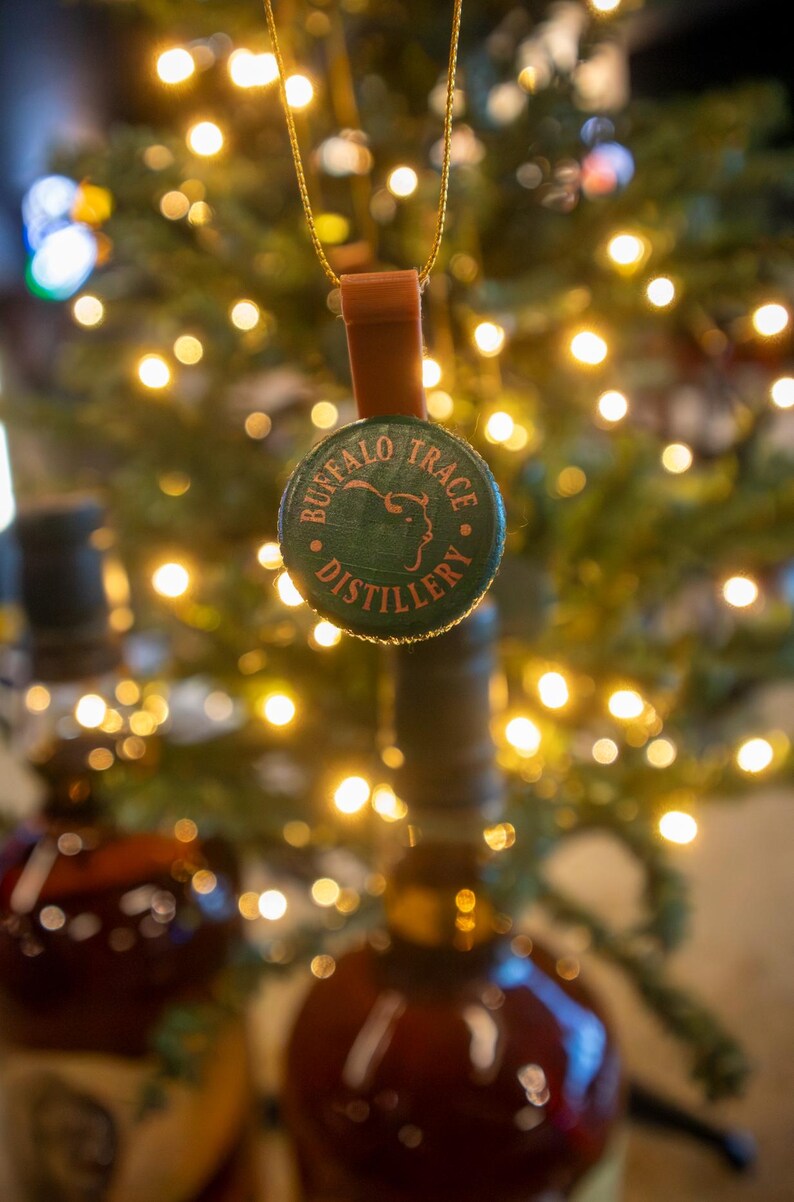 May include: A Buffalo Trace Distillery bottle cap ornament with a brown leather strap and gold-colored string. The green cap features the distillery's name and logo. The background is blurred with Christmas tree lights.