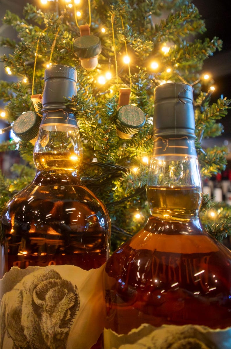 May include: Two bottles of amber-colored liquor with dark green caps, set against a backdrop of a Christmas tree with twinkling lights. One bottle features a label with a bison illustration. The scene evokes a festive, holiday spirit.