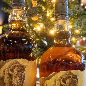 May include: Two bottles of amber-colored liquor with a label featuring a bison. The bottles have dark green caps and are set against a backdrop of a Christmas tree with twinkling lights. The labels are torn around the edges.