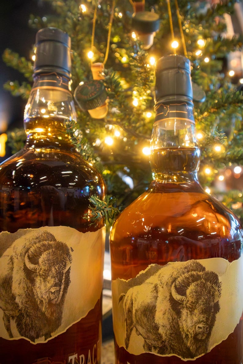 May include: Two bottles of amber-colored liquor with a label featuring a bison. The bottles have dark green caps and are set against a backdrop of a Christmas tree with twinkling lights. The labels are torn around the edges.