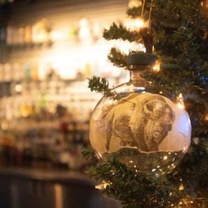 Może przedstawiać: Szklana ozdoba choinkowa w przezroczystym kolorze z nadrukowanym na przodzie vintage'owym obrazkiem bizony. Ozdoba jest zawieszona na zielonej choince z lampkami.