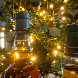 May include: Two glass bottles of amber-colored liquor with dark green caps are in the foreground. A Christmas tree with string lights and bottle cap ornaments is in the background. The bottles are partially filled.