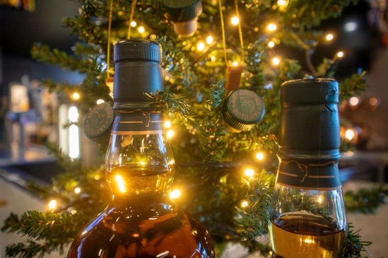 May include: Two glass bottles of amber-colored liquor with dark green caps are in the foreground. A Christmas tree with string lights and bottle cap ornaments is in the background. The bottles are partially filled.