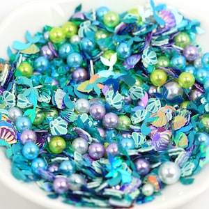 May include: A white bowl filled with a mix of mermaid-themed craft supplies. The mix includes iridescent blue, green, and purple sequins in various shapes, along with pearl beads in shades of blue, green, and purple. The sequins are shaped like shells and mermaid tails.