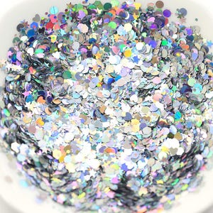 Könnte beinhalten: Eine weiße Schüssel gefüllt mit irisierendem Glitzer in verschiedenen Formen, darunter Sterne, Herzen und Kreise. Der Glitzer reflektiert mehrere Farben und erzeugt einen schimmernden Effekt. Ideal für Bastelarbeiten, Nagelkunst oder Partydekorationen.