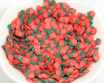 May include: A white bowl filled with red and green strawberry-shaped polymer clay slices. The strawberries have white seeds and green tops. These small, decorative pieces are likely used for crafts or nail art.