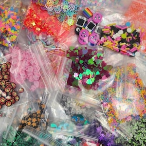 Puede incluir: Una colección de diversos suministros para manualidades, que incluye lentejuelas coloridas, confeti y botones decorativos, empaquetados en bolsas de plástico transparente. El surtido presenta una variedad de formas y colores, como estrellas, flores y caras sonrientes.