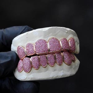 May include: A set of custom-made gold teeth grills, encrusted with pink gemstones. The grills are displayed on a white dental mold against a dark background. The gold frames are visible, showcasing the intricate design and craftsmanship.