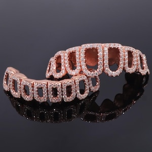 Może przedstawiać: Grillz w kolorze różowego złota z prostokątnym i owalnym wzorem, inkrustowane małymi, błyszczącymi białymi kamieniami. Grillz są prezentowane na odblaskowej czarnej powierzchni, podkreślając ich skomplikowane detale i luksusowy wygląd.
