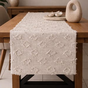 May include: A cream-coloured linen table runner with a diamond pattern and fringed edges. It sits on a wooden table with a black metal base. Decorative elements include a vase with dried plants and a stone sculpture.