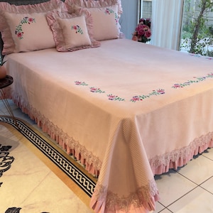 May include: A pink bedspread and pillow set with floral embroidery. The bedspread has a ribbed texture and a lace trim. The pillows have ruffled edges. The set is designed for a bed.