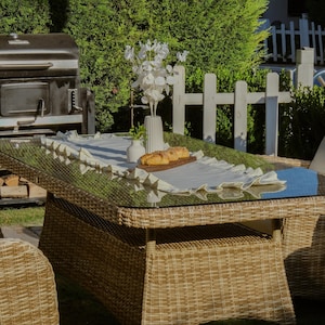 May include: Outdoor dining table with a glass top and woven wicker base. The table is set with a white runner, a vase of white flowers, and a wooden serving board with bread. A grill and white picket fence are in the background.
