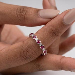 Könnte beinhalten: Ein silberfarbener Ring mit einer Reihe abwechselnder, rund geschliffener roter Rubine und kleiner, klarer weißer Diamanten in Zargenfassung. Die Edelsteine sind in einem durchgehenden Muster um den gesamten Metallring herum angeordnet.