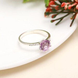 May include: A silver ring with a large, round, light purple gemstone as the focal point. The band is thin and features small, clear stones on one side. The ring is set against a white background with a hint of red and green floral accents.