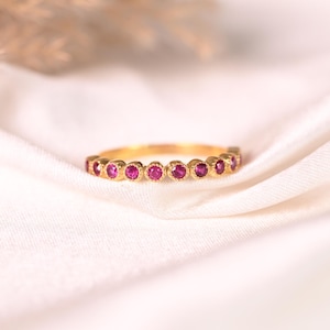 Op de afbeelding: Een gouden ring met een reeks kleine, ronde, felroze stenen in individuele zettingen. De stenen zijn in een doorlopende rij geplaatst langs de bovenste helft van de dunne, gepolijste gouden metalen band, wat zorgt voor een verfijnde uitstraling.
