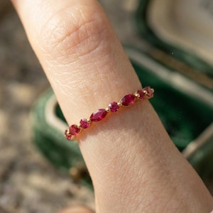 Peut inclure: Une bague en or ornée d'une série de rubis rouges alternant entre une taille ronde et une taille marquise sur le dessus. Les pierres sont disposées selon un motif répétitif, fixées par de petites griffes en or, formant une ligne continue.