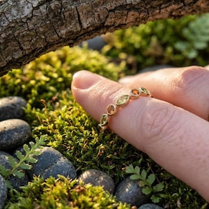 Puede incluir: Un anillo de banda dorada con una serie de piedras preciosas alternas, redondas y de corte marquesa, en tonos verdes y naranjas. Las piedras están engastadas en un delicado marco de metal con borde texturizado, creando un patrón continuo.