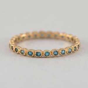 May include: A gold eternity band featuring a continuous row of small, round, blue gemstones set in a beaded metal frame. The ring has a textured, scalloped edge surrounding each individual stone, creating a uniform and detailed circular design.